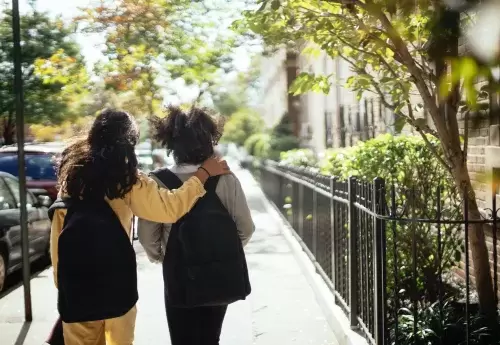 Two people walking down the street with one putting their arm over the shoulder of the other.