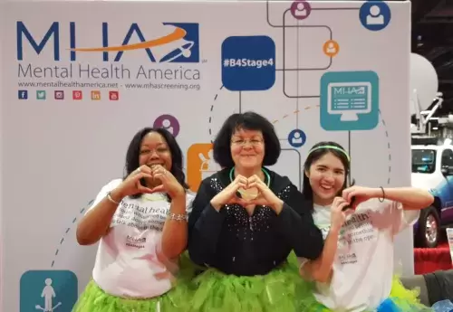 Three women in green tutus posing with their hands in the shapes of hearts in front of an MHA banner