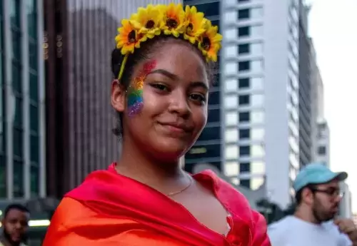 Woman with a flower crown and rainbow sparkle makeup looking at the camera with a rainbow flag wrapped around her.